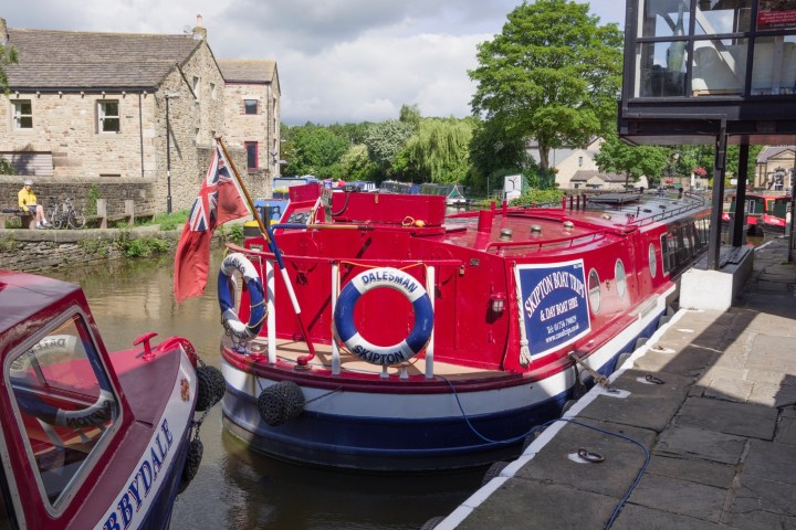 Red canal boat with lifebuoys docked by stone building and trees on a sunny day.