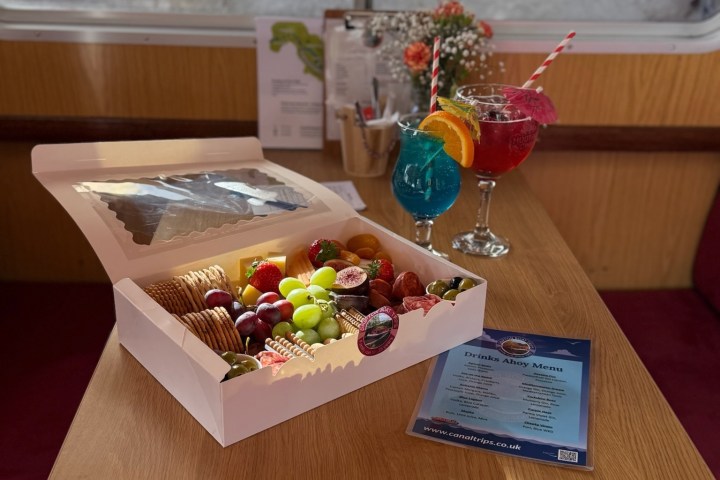 Wooden table with fruit platter and two colorful cocktails by a window.