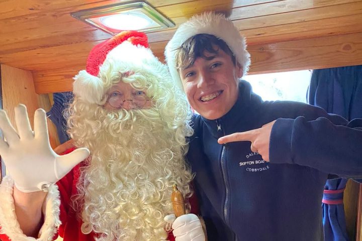 Santa and a smiling person in a festive hat posing indoors with a bell.