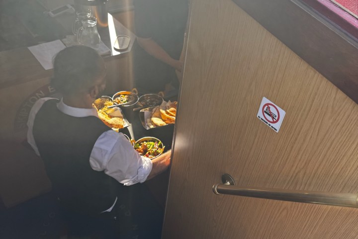 Man in vest carrying food trays down narrow stairs with caution sign.