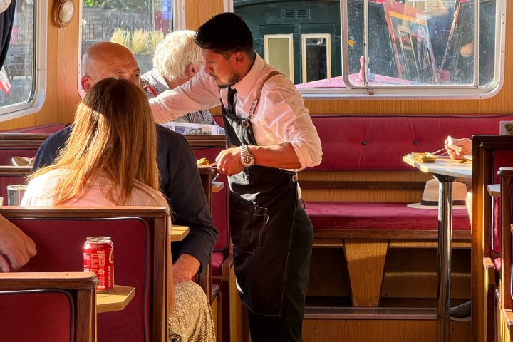 Waiter serves customers on a boat with wooden interiors and red seating.