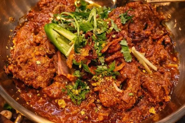 Spiced curry in a brass pot, garnished with herbs and a spoon placed inside.